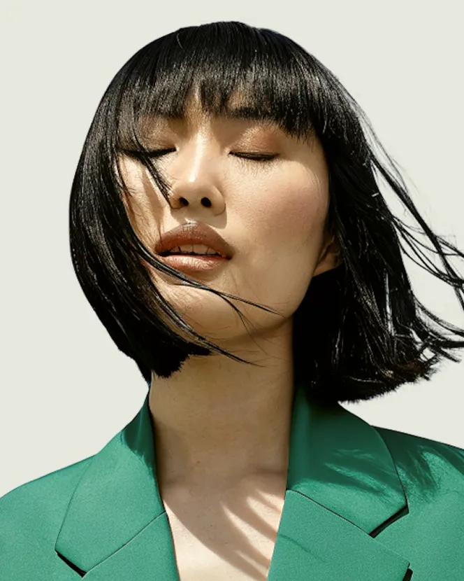 Woman with black bob haircut and bangs wearing a green blazer, eyes closed and hair blowing.